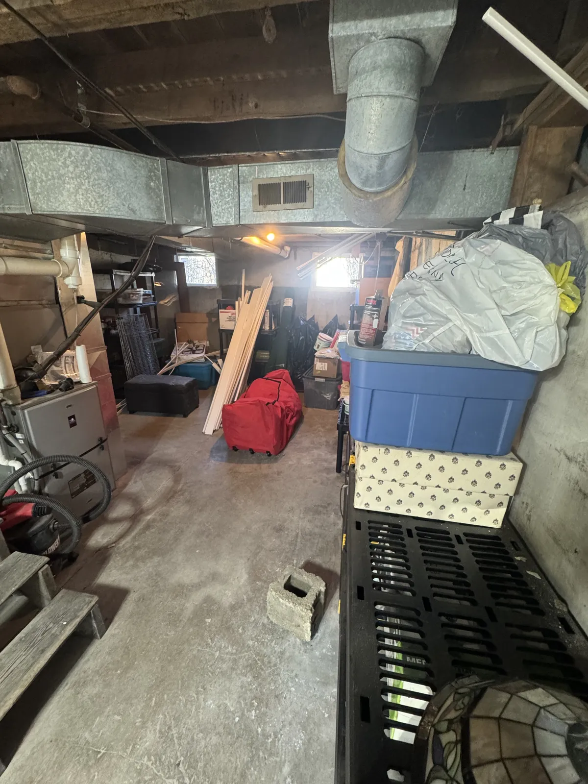 Unfinished basement with ductwork, furnace, bins, red duffel, lumber, boxes and cinder block