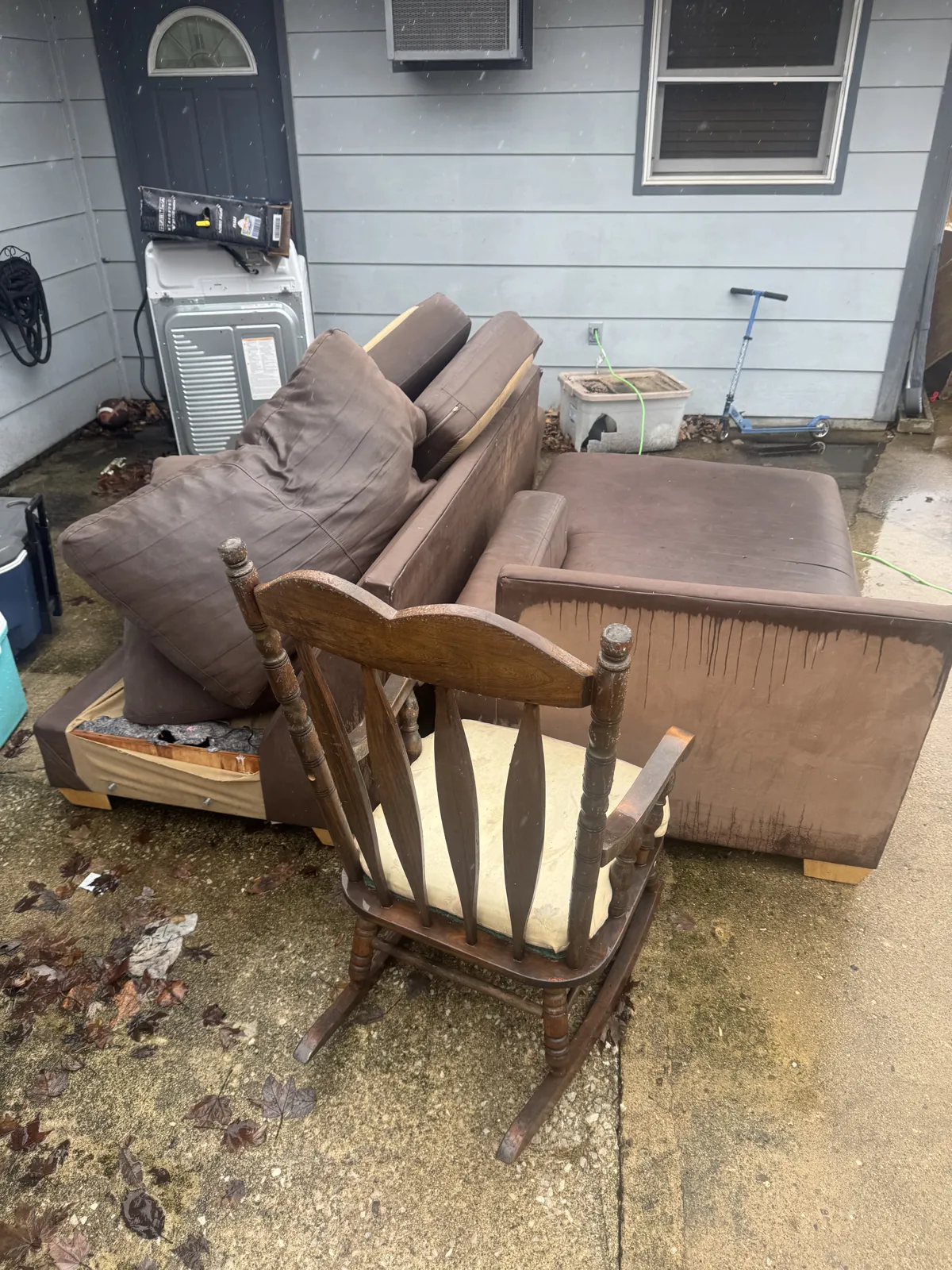 Brown sectional couch pieces and a wooden rocking chair on a wet concrete patio beside blue siding