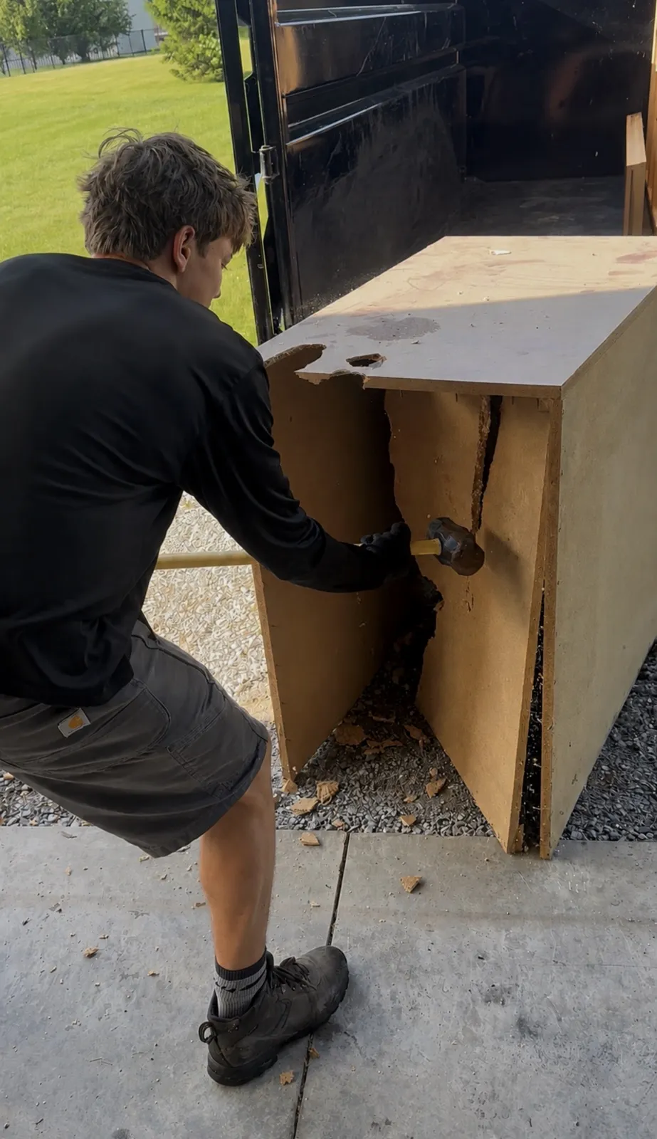 Crew member in black shirt and Carhartt shorts breaking apart a particleboard cabinet with a sledgehammer
