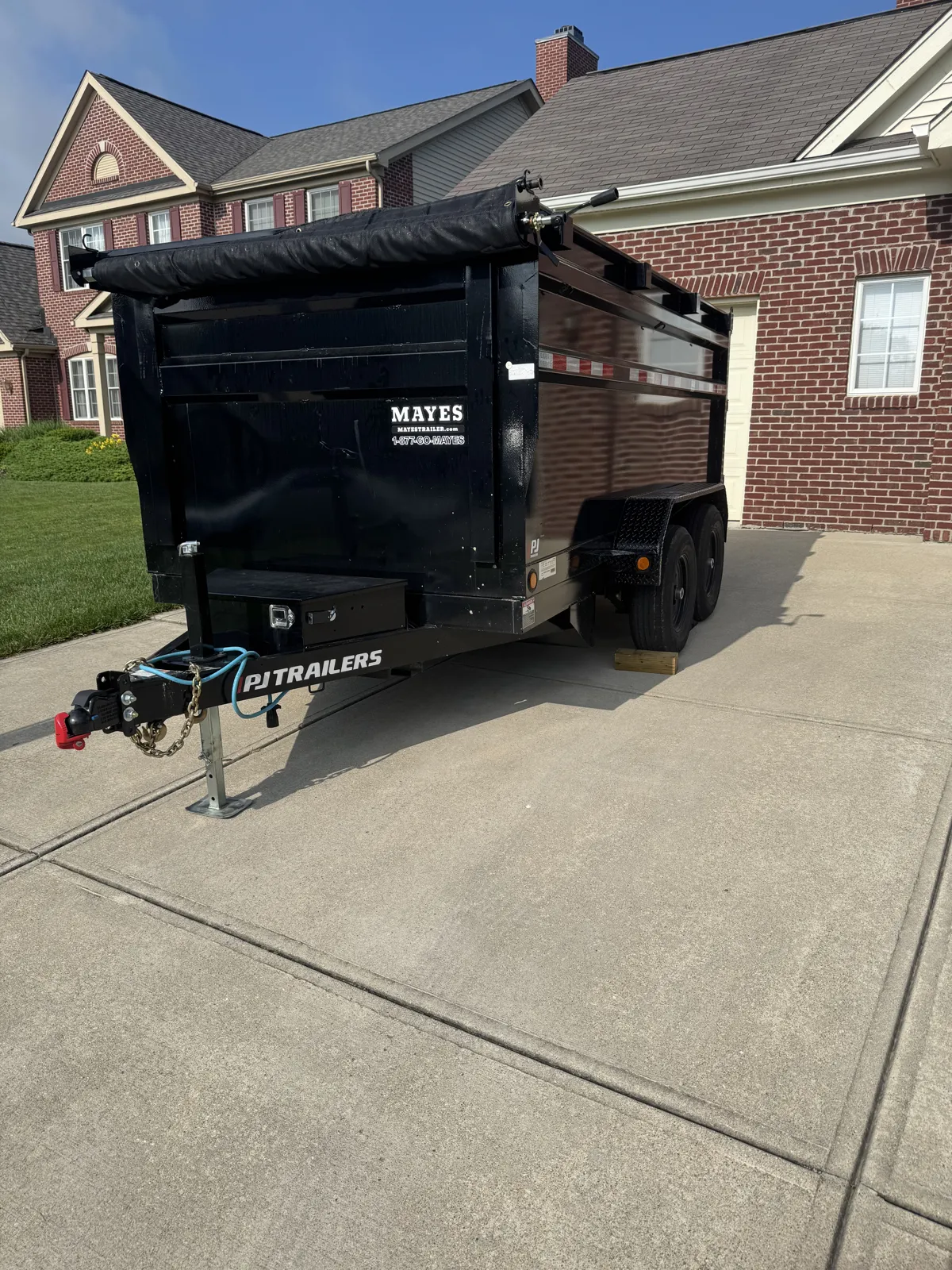 Black PJ dump trailer (tarp rolled) parked on a driveway in front of brick homes, blue sky