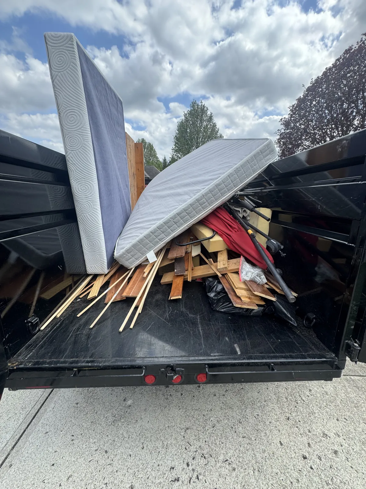 Open dump trailer loaded with a mattress, box spring, lumber and red umbrella under a cloudy blue sky