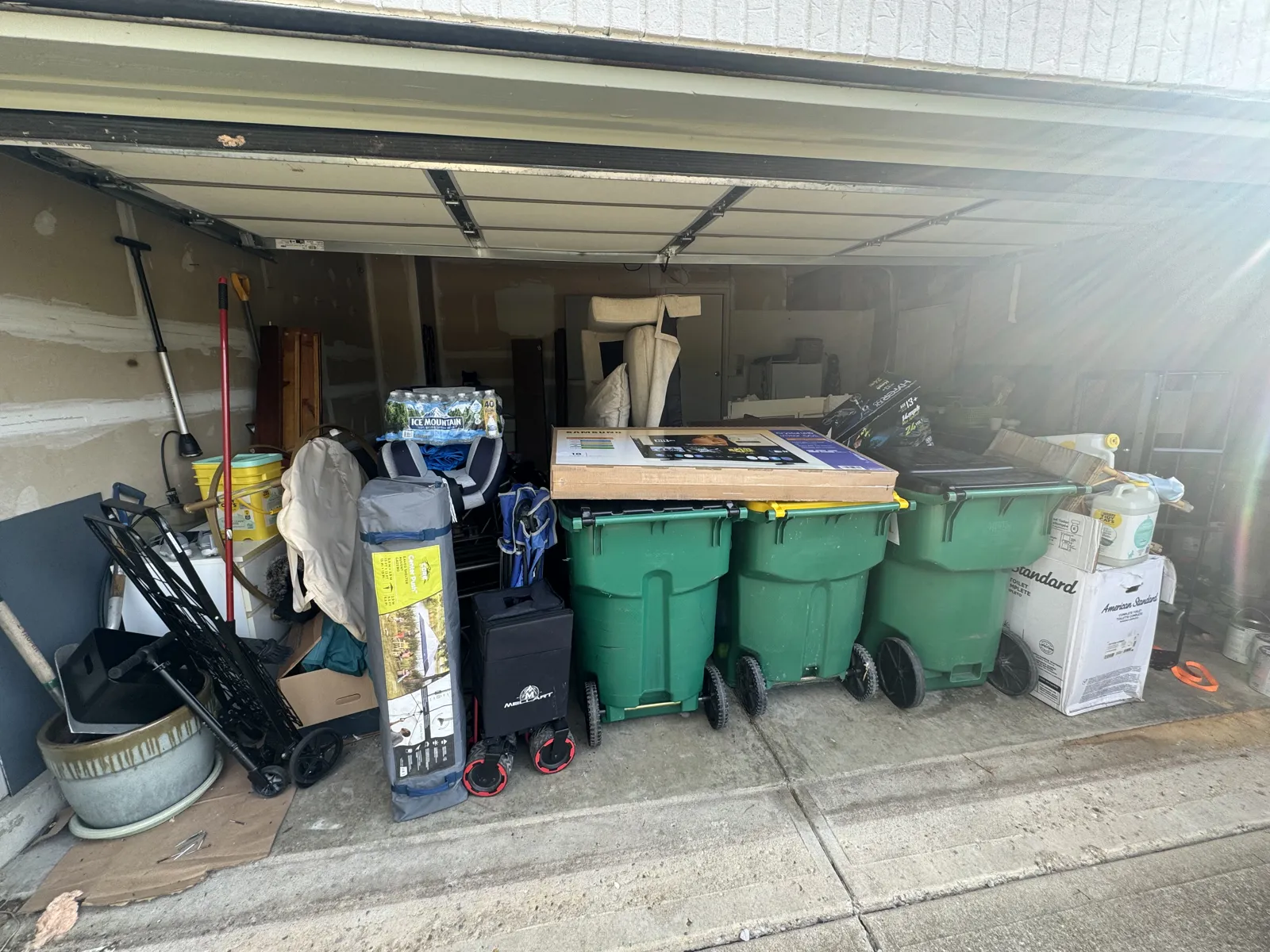 A garage bay filled with three green trash carts, a boxed TV, camping gear, dolly and boxes