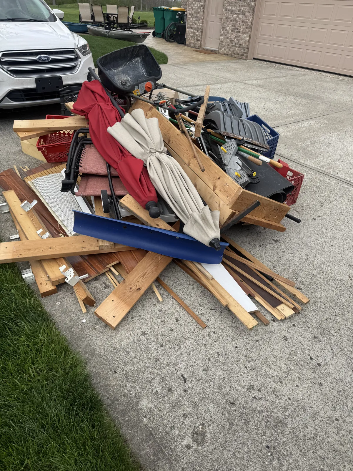 Driveway pile of lumber, patio umbrellas, wheelbarrow, milk crates, snow shovel and tools next to a white