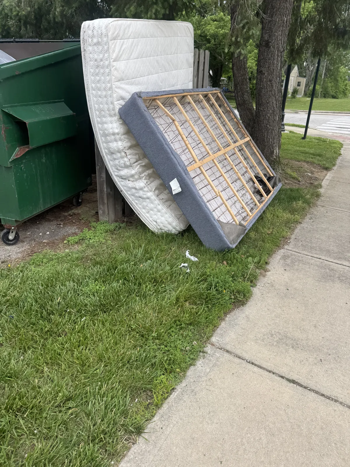 Mattress and box spring leaning against a fence beside a green dumpster, next to a sidewalk