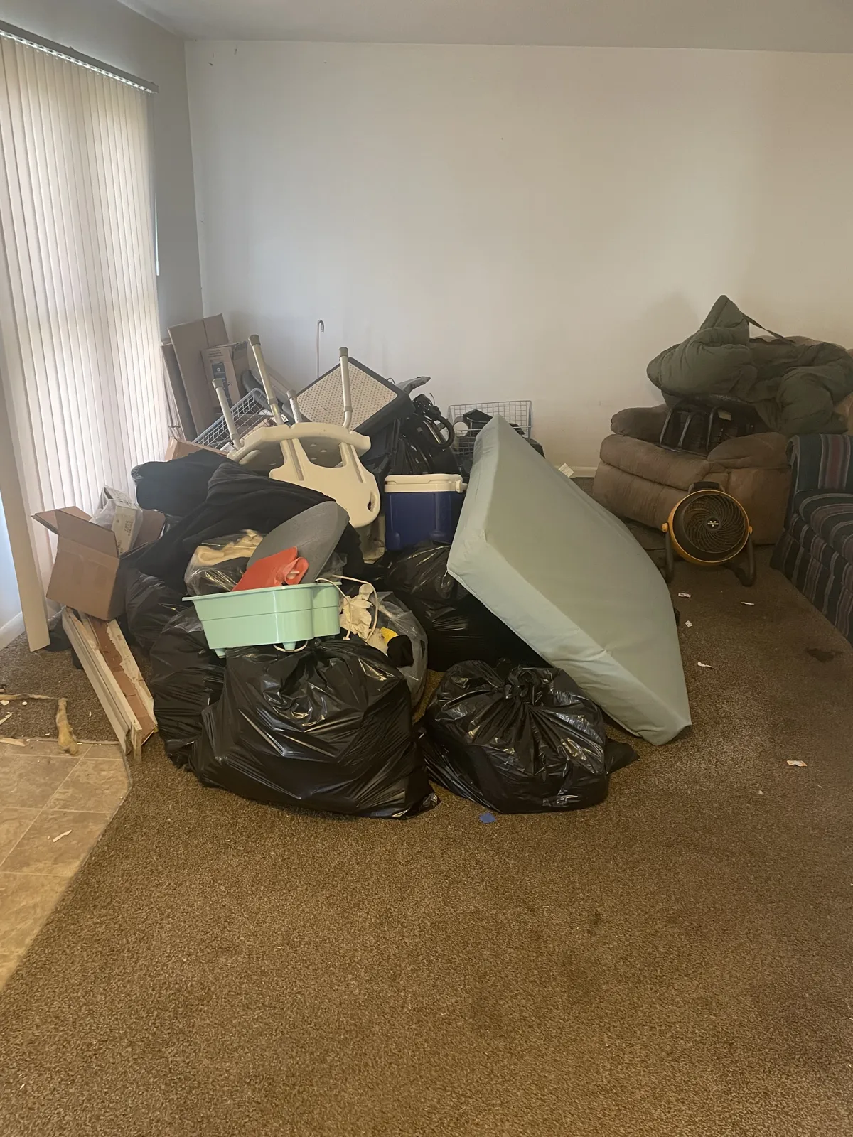 Carpeted living room with a large pile of black trash bags, a mattress, cooler, broken chair and boxes