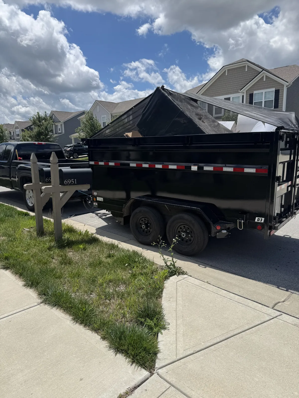 Loaded, tarped black dump trailer hitched to a black pickup at the curb in a suburban neighborhood, blue sky